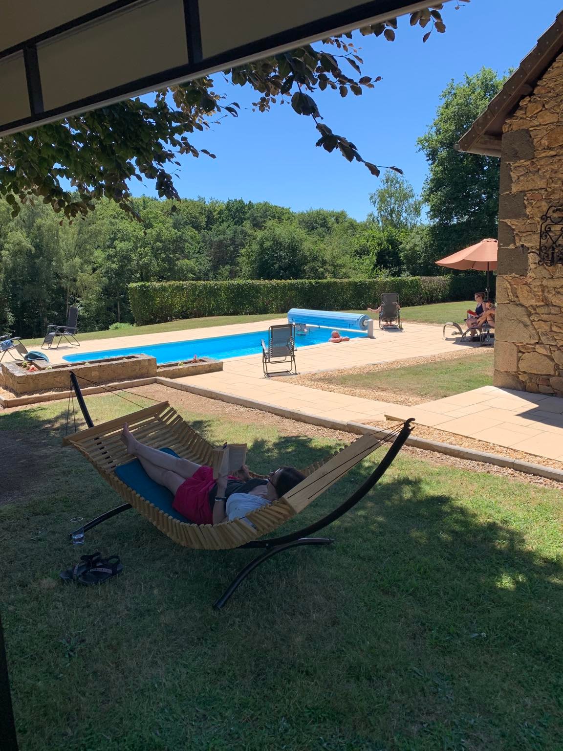 Person relaxing in a hammock by a pool with a stone building and trees in the background