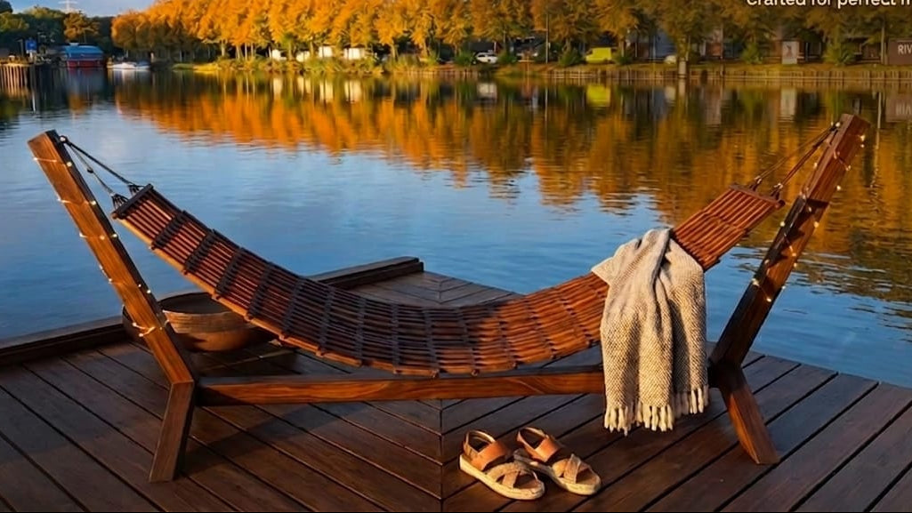 Wooden hammock on a deck overlooking a calm lake with trees and a clear sky.